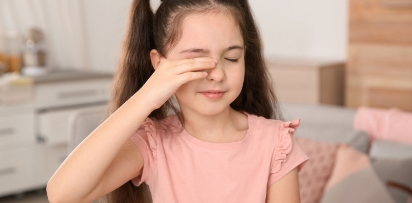 A young girl wearing a pink t-shirt, rubbing her right eye with her hand