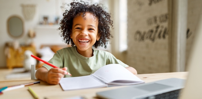 A young boy smiling at the camera, sat at the table doing work in a school book