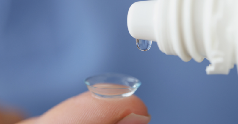 A close-up of a contact lens on the end of a person’s finger with a drop of contact lens solution hovering over it.