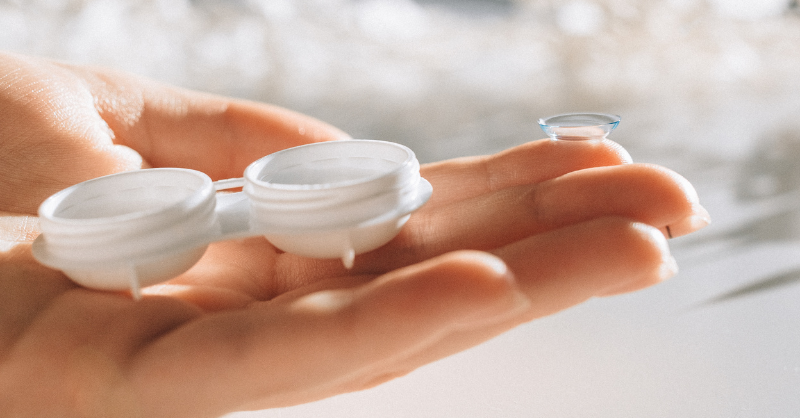 An outstretched hand with a contact lens storage pot in the palm of the hand and a single contact lens on the tip of a forefinger.