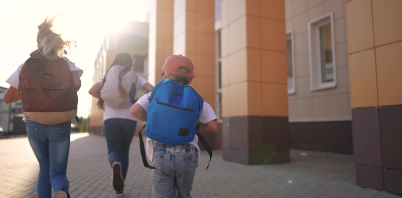 A group of young children wearing backpacks, running towards a building